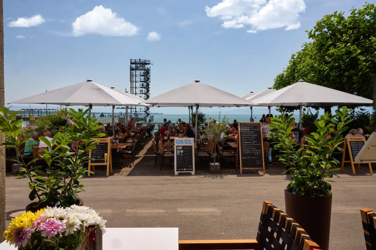 Das Delphi in Friedrichshafen mit Weißen Sonnenschirmen im Gastronomiebetrieb und Aussichtsturm im Hintergrund.